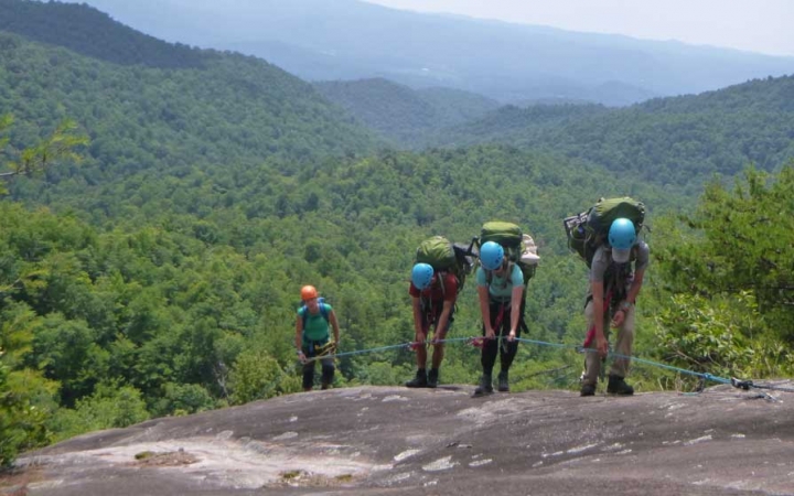 Four students wearing helmets and backpacks hold on to a rope as they scramble up a rock incline. Behind them is a mountainous landscape covered in trees.